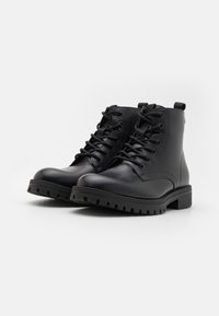 Botas de cuero negro con punteras redondeadas, suelas de goma texturizadas y ojales de metal para los cordones. Cuentan con una pestaña para facilitar el uso.