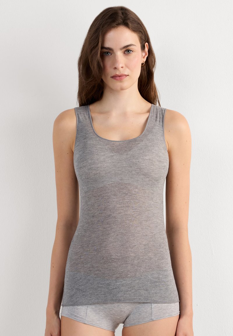 Frau mit langen braunen Haaren, die ein eng anliegendes graues ärmelloses Tanktop und passende graue Shorts trägt, steht vor einer schlichten weißen Wand.