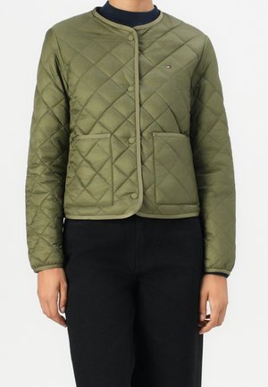 Person iført olivengrøn quiltet jakke med trykknapper og to frontlommer, kombineret med sorte bukser og en mørk undertrøje.