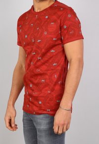 Man i röd T-shirt med grå racing-tema ikoner och ljusgrå jeans, som står mot en vanlig ljus bakgrund.