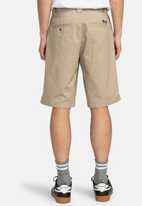 Pantaloni corti khaki a taglio dritto, con due tasche posteriori e un'etichetta con il marchio. Indossati con scarpe da ginnastica nere e bianche e calze grigie a righe.