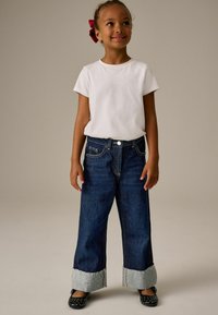 I jeans in denim presentano un colore blu scuro, gamba larga e orli sfilacciati di un blu chiaro. Abbinati a una t-shirt bianca e scarpe nere a pois.