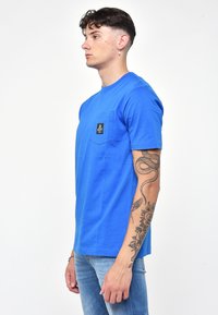 Maglietta blu a maniche corte in cotone con tasca e un'etichetta nera. Il modello indossa jeans in denim chiaro, mostrando i tatuaggi sull'avambraccio.