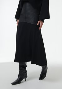 Falda negra midi con una cinturilla de cuero acanalado, combinada con botas negras de punta afilada hasta la rodilla. Textura suave, diseño fluido.