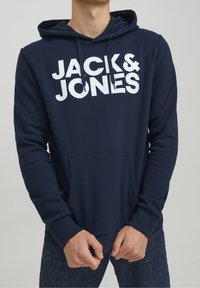 Námořnická modrá mikina s kapucí vyrobená z bavlněné směsi, s přední kapsou, šňůrkovou kapucí a odvážným bílým logem "JACK & JONES" na hrudi.