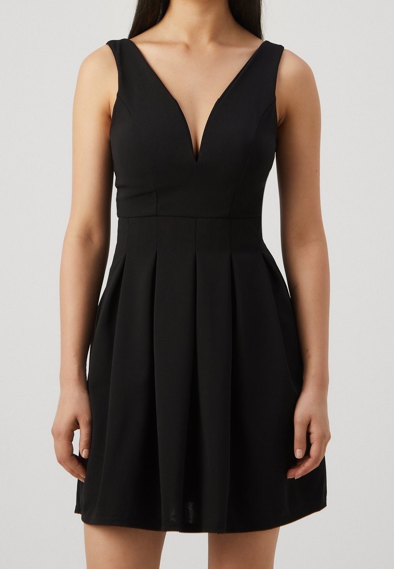 Robe noire sans manches avec un décolleté en V profond et une jupe plissée portée par une personne sur un fond uni.