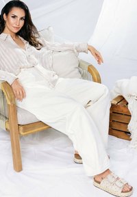 Helles gestreiftes Hemd, lockere weiße Hose und beigen Sandalen mit dekorativen Schnallen. Sitzt auf einem Holzstuhl mit Kissen.