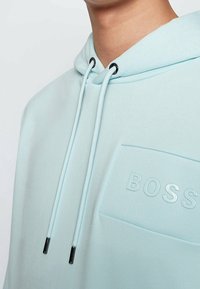 Sudadera de color azul claro con un bolsillo texturizado en el pecho que presenta la palabra "BOSS" en letras en relieve, cordones blancos con puntas negras.