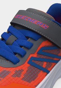 Κοντινό πλάνο πορτοκαλί και γκρι αθλητικού παπουτσιού Skechers με μπλε κορδόνια και γκρι λουράκι με το λογότυπο Skechers σε κόκκινο και μπλε χρώμα.