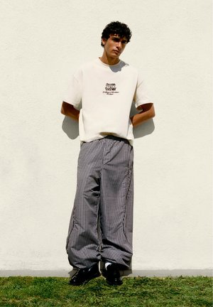 Mladý muž s kučeravými vlasmi stojí pri bielej stene, má na sebe biele tričko s grafikou, kárované široké nohavice a čierne topánky na tráve.