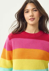 Pull en tricot multicolore avec de larges rayures horizontales rouges, roses, orange, jaunes et bleu clair. Encolure ronde et texture côtelée.