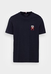 T-paita tummansinisestä puuvillasta, jossa on lyhyet hihat ja pieni punavalkoinen kirjailtu logo vasemmalla rintakehässä. Klassinen pyöreä pääntie.