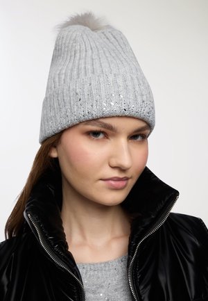 Donna che indossa un berretto di maglia grigio chiaro con un pom-pom e paillettes, un maglione coordinato e un cappotto imbottito nero lucido con la zip aperta.