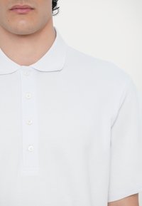 Hvit polo skjorte laget av teksturert bomull med klassisk krage, fem-knappslukking og korte ermer. Glatt stoff med minimalt med detaljer.