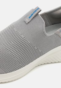Scarpa slip-on grigia in maglia con tomaia a texture, dotata di una suola in gomma bianca e un accento blu vicino al tallone per una facile vestibilità.