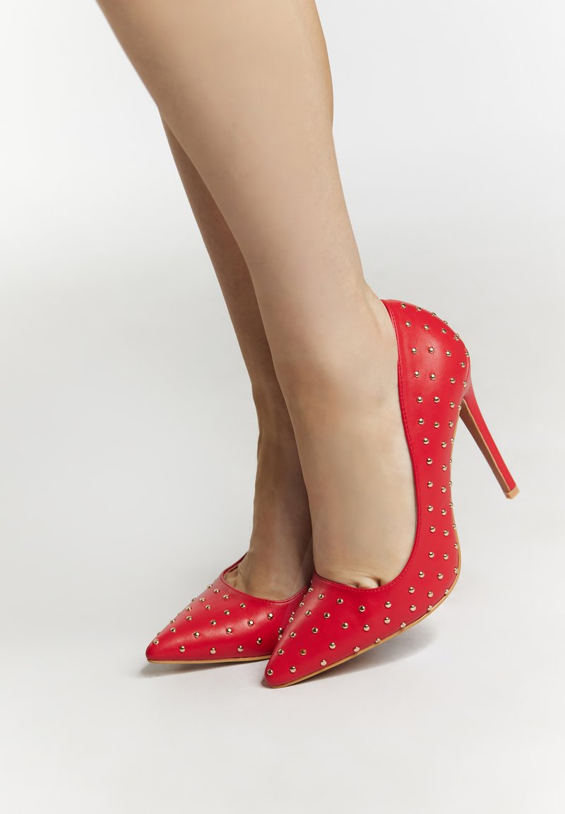 Scarpe rosse in pelle con tacco alto e punta affilata, caratterizzate da borchie dorate disposte in un motivo lungo i lati e un sottile tacco a stiletto.