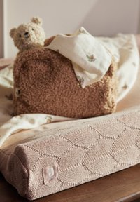 Een gevuld teddybeer in een gestructureerd bruin pluche bed met een witte bloemenpatroon deken, geplaatst op een roze gebreid matras met een geometrisch ontwerp.