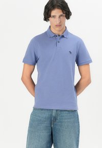 Fialové polo tričko vyrobené z bavlny, s malým logem na levé straně hrudi, krátkými rukávy a zapínáním na tři knoflíky. Nosené s modrými džínami.