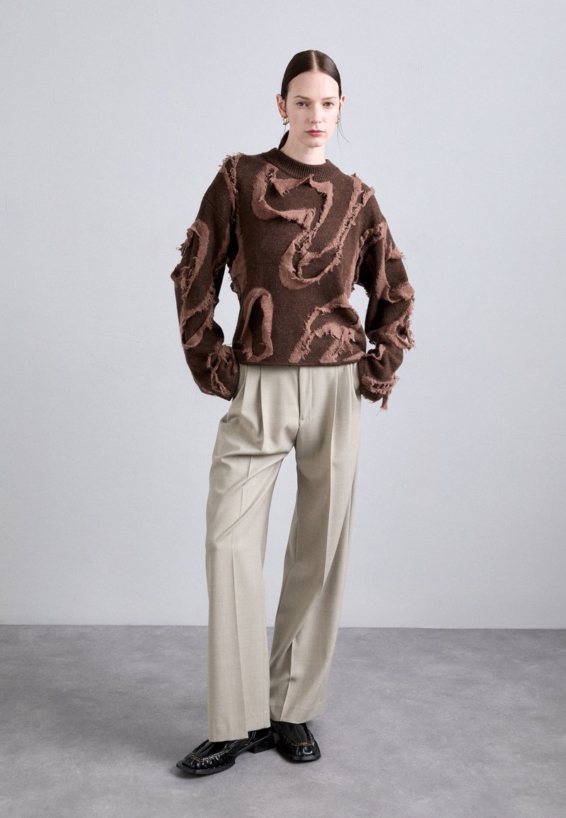 Bruine gebreide trui met rafelige accenten, gecombineerd met lichtbeige plisséadbroeken en zwarte schoenen. Zachte texturen en contrasterende kleuren.