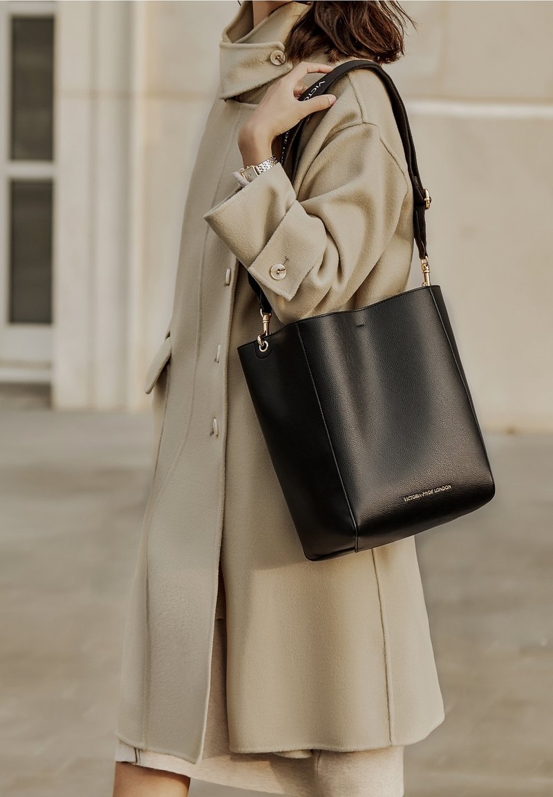 Femme portant un manteau beige porte un sac fourre-tout en cuir noir avec le logo "Victoria Hyde London" à l'extérieur près d'un bâtiment.