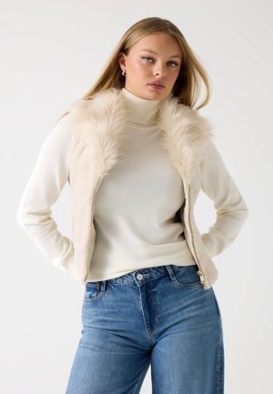 Gilet in pelliccia finta di colore crema, con chiusura a zip, abbinato a un dolcevita chiaro e jeans blu a vita alta. Texture morbida, vestibilità casual.