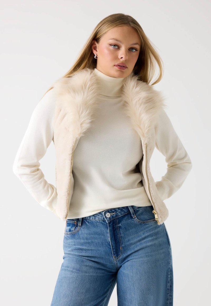 Gilet in pelliccia finta di colore crema, con chiusura a zip, abbinato a un dolcevita chiaro e jeans blu a vita alta. Texture morbida, vestibilità casual.
