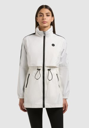 Mujer con chaqueta blanca larga con cremallera, cuello alto, cremallera negra y cordones, y pantalones negros, de frente sobre fondo liso.