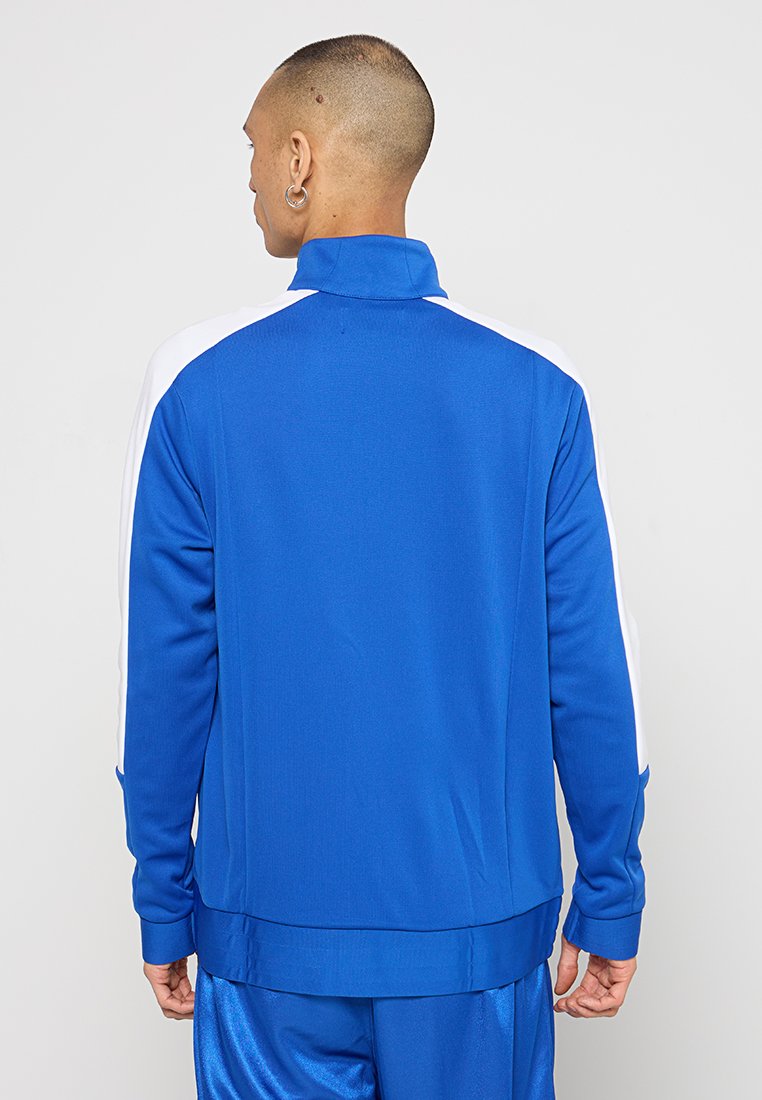Blue athletic jacket with a high collar, white side accents, made of smooth fabric. Features a relaxed fit and long sleeves.