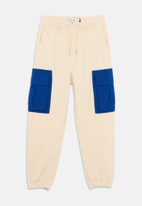 Beige bomull joggebukser med elastisk midje, med to store blå cargo-lommer på lårene. Tømmes ved anklene.