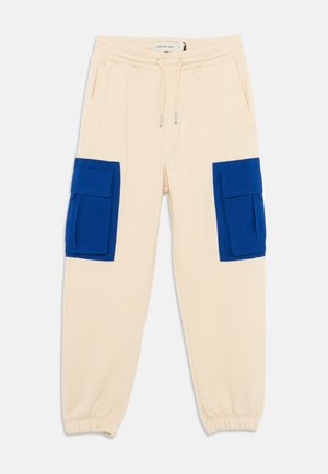 Beige cotton sweatpants with elastic waistband, featuring two large blue cargo pockets on the thighs. Cuffed at the ankles.