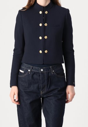 Kvinde iført en mørkeblå cropped jakke med gyldne knapper og mørkeblå jeans, vist fra skuldre til hofter mod en ensfarvet baggrund.