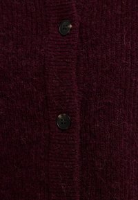 Vyno raudonos spalvos megztas kardiganas su šiek tiek tekstūruota paviršiaus, ribinėmis kraštinėmis ir dviem juodais mygtukais priekyje.