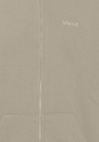 Felpa con zip color beige caratterizzata da una texture morbida, tasche frontali e un logo di colore chiaro nella zona sinistra del petto.