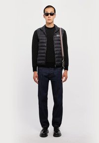 Gilet rembourré noir sur un pull en tricot noir texturé, jean sombre et chaussures noires. Lunettes de soleil et sac avec une bandoulière rayée.