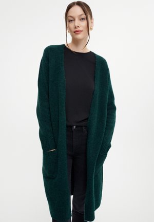 Cardigan verde lavorato a maglia con apertura frontale, maniche lunghe e tasche. Presenta una texture morbida e polsini a coste, indossato sopra una maglietta nera e jeans.