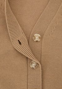 Dzeltenbrūns adīts kardigans ar ribotu apkakli, kurā ir divi lieli, apaļi, gaiši krāsas pogas un maza caurumiņa poga sānos.