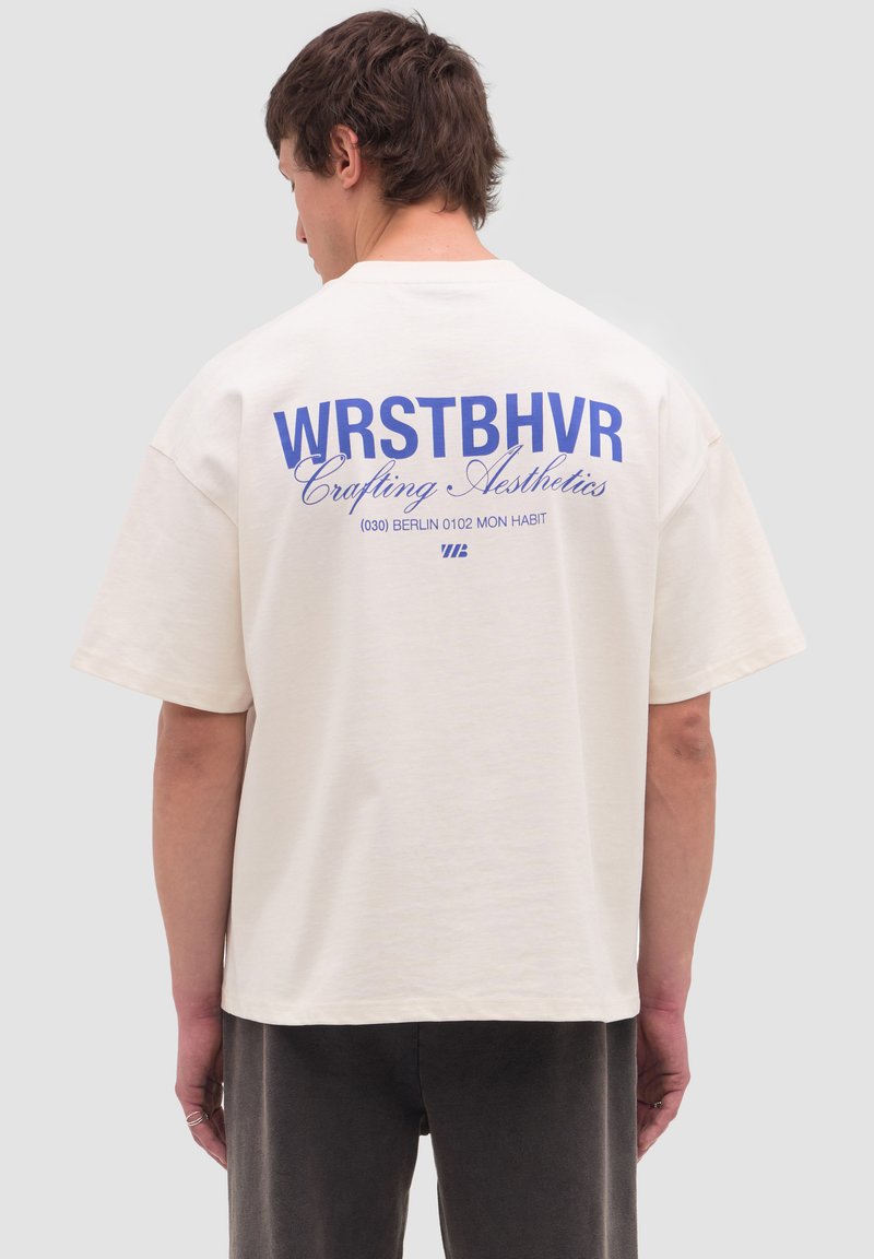 Mies selinpäin, yllään valkoinen ylisuurikokoinen t-paita, jossa sininen teksti "WRSTBHVR Crafting Aesthetics" selässä.
