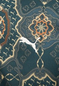 Krāsains audums ar sarežģītām ziedu un ģeometriskām šķautnēm tumši zaļā, krēmkrāsas un oranžā krāsā, ar balto Puma logotipu.