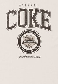 Cremefarvet t-shirt med fed, slidt "ATLANTA COKE" tekst, et cirkulært Coca-Cola logo og sloganet "Du kan ikke slå følelsen!"