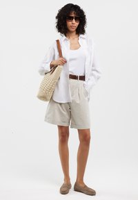 Pantalones cortos de algodón beige, camiseta sin mangas blanca, camisa blanca de botón de gran tamaño, cinturón marrón, bolsa grande de mimbre y mocasines marrones; gafas de sol oscuras.