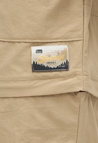 Μπεζ ύφασμα με ορθογώνια ραμμένη ετικέτα Columbia Sportswear Co. που απεικονίζει δέντρα και κείμενο "Δοκιμασμένο Ανθεκτικό" και "Από το 1938."