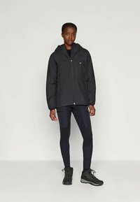Veste imperméable noire avec une fermeture éclair à l'avant, une capuche et des poches latérales, associée à des leggings sombres ajustés et de solides bottes de randonnée noires.