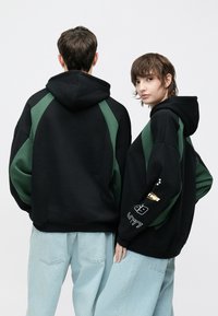 Must kapuutsiga dressipluus roheliste detailidega, suur lõige, kapuutsi kinnituspael, varrukatel graafilised trükid. Kandatud hele siniste teksadega.