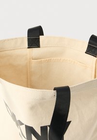 Plátená taška na rameno s čiernymi rukoväťami, béžovým vnútrom a vreckom; na jednej strane výrazný čierny nápis. Čistý dizajn, pevná konštrukcia.