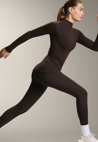 Top atletico marrone scuro con colletto alto e cerniera, abbinato a leggings coordinati. Il tessuto appare elastico e strutturato, adatto per l'esercizio.