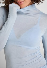 Kvinna som bär en genomskinlig ljusblå långärmad topp och lyfter kragen vid munnen, vilket visar en matchande blå cami under.