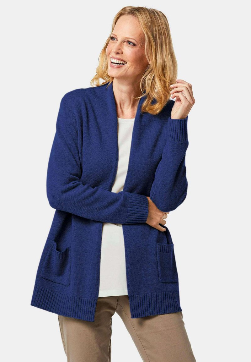 Cardigan en tricot bleu foncé avec col châle, poignets côtelés et deux poches avant. Texture douce et coupe décontractée, porté sur un top blanc.