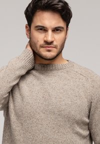 Jasnobrązowy sweter z dzianiny z okrągłym dekoltem i prążkowanymi mankietami. Tekstura jest miękka, z delikatnym wzorem w drobne speckles. Model dotyka szyi.