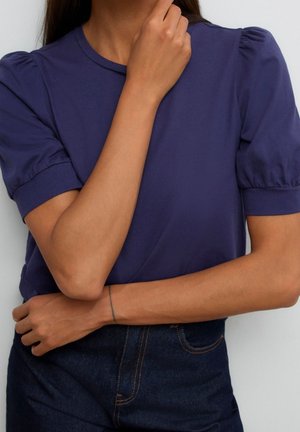 Donna che indossa una t-shirt blu navy con maniche a sbuffo e jeans blu scuro, con una mano che tocca il mento e l'altra incrociata sulla vita.