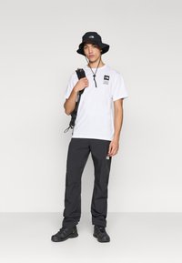White short-sleeve t-shirt with logo patch, paired with black waterproof pants and black bucket hat. Black hiking shoes complete the look.